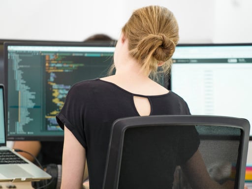 A woman facing away from the camera, sitting at a computer, working on some code. 